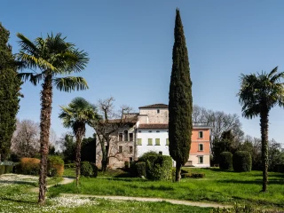 Esterno del museo Archeologico ripreso dal giardino. Edificio composto da più corpi affiancati, con una parte in mattoni, una sezione centrale bianca con imposte verdi e una parte arancione sulla destra. Davanti all’edificio si trovano un prato verde, aiuole e varie piante, tra cui palme e un alto cipresso. Sullo sfondo sono visibili alberi spogli e un cielo limpido.