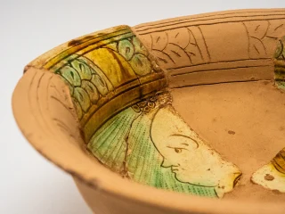 Scodella in ceramica graffita ricomposta e reintegrata. I frammenti originali presentano motivi verdi e gialli e parte del profilo di un volto stilizzato. Il resto della superficie restaurata è liscia e priva di decorazioni. Epoca rinascimentale.
