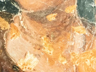 Dettaglio ravvicinato di un affresco seicentesco deteriorato che mostra la parte superiore del busto e del collo di una figura femminile. La superficie pittorica presenta ampie aree di picchiettatura che interrompono la continuità del disegno originale. Le pennellate evidenziano tonalità rosate e brune sul volto e sul collo, con frammenti di fondo scuro ai lati.