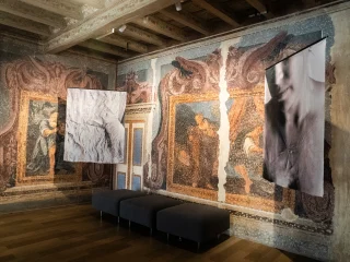Interno di una sala con pareti decorate da grandi affreschi seicenteschi dai toni blu, arancioni e rosati, raffiguranti figure umane incorniciate da motivi ornamentali. Dal soffitto ligneo decorato scendono due grandi stampe fotografiche in bianco e nero, sospese al centro della stanza della fotografa Olivia Arthur esposte in occasione di una mostra temporanea. Sotto le stampe sono collocati tre pouf scuri allineati. Il pavimento è in legno chiaro.
