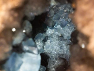La foto in primo piano di un geode rivela cristalli sfaccettati di un azzurro brillante. I cristalli scintillanti sono circondati da roccia ruvida e marrone chiaro. La luce illumina l'interno della cavità, rivelando cristalli di diverse dimensioni, altamente addensati.