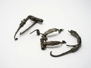 Gruppo di fibule in argento risalenti al primo secolo a.C. probabilmente di origine celtica. Presentano decorazioni plastiche con globetti e spirali e grandi molle attorcigliate. Gli oggetti sono disposti su una superficie bianca e appaiono frammentati o deformati.