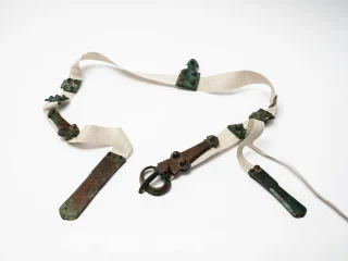 Antica cintura longobarda composta da decorazioni originali in bronzo applicate su una lunga striscia in tessuto chiaro. Al centro è presente una fibbia che connetteva le due estremità della cintura. Alle estremità si trovano ulteriori placche allungate in bronzo.