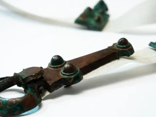 Dettaglio ravvicinato di una placca di cintura in bronzo di epoca longobarda. Sulla sinistra è visibile il gancio che connetteva le due estremità della cintura. Sono presenti tre borchie decorative. Sullo sfondo è bianco sono visibili altri elementi decorativi in bronzo.