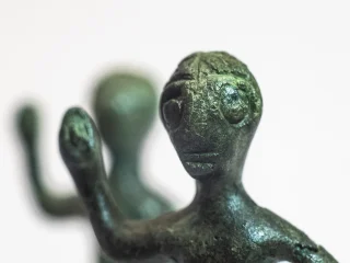 Primo piano di una piccola statuetta antropomorfa in bronzo di colore verde, con superficie irregolare e tratti del volto incisi. La figura ha un braccio sollevato. Si tratta della rappresentazione stilizzata di un guerriero nell'atto di reggere una lancia. Sullo sfondo, sfocata, è visibile un’altra statuetta simile. Questi guerrieri venivano offerti in contesti votivi.