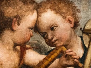 Dettaglio ravvicinato della pala di San Gottardo del Pordenone che raffigura due putti con capelli ricci, testa contro testa. Una delle figure tiene tra le mani un piccolo strumento musicale a fiato. La superficie pittorica presenta una trama visibile.