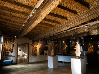 Interno di una sala museale del Ricchieri con soffitto ligneo decorato da motivi geometrici e floreali. La stanza espone diverse sculture, alcune delle quali accolte in teche trasparenti su piedistalli grigi e altre opere lungo le pareti. Sulla sinistra si vede una finestra e una porta che conduce a un altro ambiente illuminato. Il pavimento è in legno chiaro.