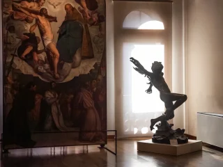 Interno del salone centrale al primo piano del museo Ricchieri, con una grande pala d'altare a sinistra rappresentante la Santissima Trinità e una scultura in bronzo a destra del volo di Icaro. La scultura è illuminata dalla luce proveniente da una porta-finestra sullo sfondo, creando una silhouette marcata. Il pavimento è in legno e il soffitto presenta travi a vista.