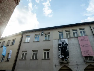 Facciata di un edificio a più piani con finestre rettangolari e due grandi banner verticali appesi: uno con una fotografia in bianco e nero e uno rosa con testo stampato. Accanto, sulla sinistra, sono appese altre bandiere verticali. In alto si vede una porzione di cielo con nuvole.