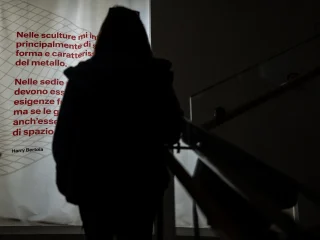 Silhouette di una persona in controluce che sale una scala interna, vista di spalle. Davanti alla persona è visibile un grande pannello illuminato con testo rosso su sfondo chiaro. A destra si intravede la ringhiera della scala.