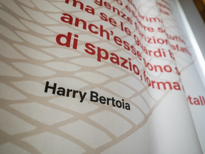 Dettaglio ravvicinato di un pannello espositivo, stampato con motivo geometrico beige sullo sfondo e testo rosso di grandi dimensioni. In basso al centro compare la scritta ‘Harry Bertoia’ in caratteri neri.