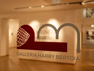 Vetro d’ingresso con il logo e la scritta ‘Galleria Harry Bertoia’; oltre il vetro si intravede uno spazio espositivo con pavimento in legno, pareti chiare e illuminazione soffusa.