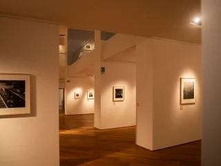 Sala espositiva con pareti bianche e pavimento in legno, illuminata da faretti a soffitto. Diversi quadri incorniciati in bianco e nero sono appesi alle pareti, ciascuno con una piccola etichetta sottostante. Le pareti creano passaggi e scorci prospettici che conducono verso altri ambienti della mostra.