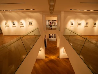 Ampio spazio espositivo a due livelli, con pavimento in legno chiaro e corridoi laterali delimitati da parapetti in vetro. Sulle pareti bianche del livello superiore sono appese fotografie incorniciate, illuminate da faretti a soffitto. In fondo, al centro, si apre una vista su un’altra sala espositiva con ulteriori immagini.