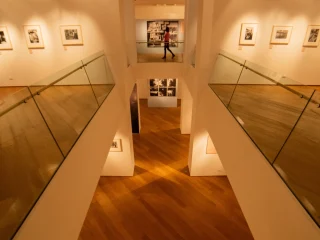 Interno di una sala espositiva a due livelli con corridoi laterali dotati di parapetti in vetro. Le pareti bianche del livello superiore sono decorate con fotografie incorniciate illuminate da faretti a soffitto. Al centro si apre un vano che permette la vista verso il piano inferiore e, sul fondo, un’altra sala con ulteriori opere esposte.