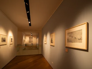 Corridoio espositivo illuminato con luci a soffitto, con stampe incorniciate appese lungo entrambe le pareti. In fondo al corridoio è visibile una balaustra in vetro che affaccia su un livello inferiore. Il pavimento è in legno e le pareti sono dipinte in toni chiari.