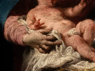 Un dettaglio di un dipinto a olio di Giambattista Pittoni che ritrae Gesù bambino in braccio alla Madonna. La pelle del bambino ha un incarnato rosato e presenta una craquelure visibile, che suggerisce la vetustà dell'opera. Il bambino è avvolto in un panno bianco drappeggiato e poggia su un tessuto rosso-brunastro. La mano di Maria lo sorregge con delicatezza. Lo sfondo è scuro e indistinto, concentrando l'attenzione sul bambino e sulla mano. Lo stile è realistico e ricco di dettagli materici.