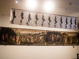 Parete espositiva con due opere sovrapposte. In alto una lunga sequenza fotografica stampata su seta della fotografa Olivia Arthur in esposizione temporanea. E' raffigurata su sfondo chiaro, in sequenza fotografica, una persona con protesi alle gambe, vestita di scuro, nell'atto di camminare. Sotto un grande dipinto murale orizzontale (680x108cm) che raffigura un ballo campestre. Realizzato nel XVI sec. da artista di scuola pordenoniana.