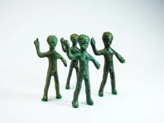 Quattro piccole statuette antropomorfe in bronzo stilizzate, disposte in gruppo su uno sfondo chiaro. Le figure hanno corpi allungati, superfici irregolari e un braccio sollevato. Si tratta della rappresentazione stilizzata di guerrieri nell'atto di reggere una lancia. Due statuette sono in primo piano e due leggermente arretrate. Questi guerrieri venivano offerti in contesti votivi.