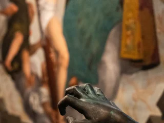 Primo piano della mano di Icaro, scultura di Luigi De Paoli. Sullo sfondo sfocata si vede la grande pala d'altare rappresentante la Santa Trinità di Gasparo Narvesa. L'effetto visivo suggerisce il tentativo di Icaro di togliere il chiodo dalla croce del Cristo.