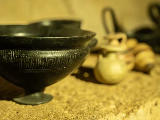 Primo piano di vasi di epoca etrusca all'interno di una tomba ricostruita. In primo piano una ciotola nera in bucchero a piede rialzato con incisioni verticali e orizzontali. Sullo sfondo, sfocati, sono presenti piccoli vasi chiari decorati con bande e altri contenitori in bucchero. L’illuminazione calda mette in risalto le texture delle superfici.