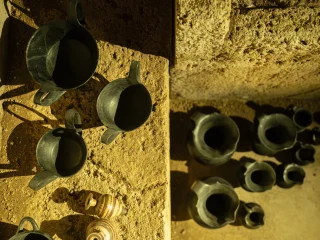 Ripresa dall'alto di vasi di epoca etrusca all'interno di una tomba ricostruita. Su un piano rialzato sono disposti tre kantharos in bucchero e due vasetti chiari decorati da bande marroni. Sul fondo è visibile una serie di brocche in bucchero. La luce laterale crea forti contrasti, evidenziando la texture ruvida delle pareti e dei contenitori.