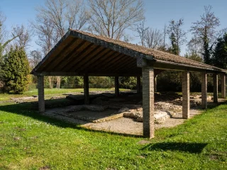Area archeologica all’aperto con un ampio tetto a falde sostenuto da pilastri in mattoni. Sotto la struttura si trovano resti di mura e pavimentazioni antiche in pietra e mattoni riferibili a una villa romana del primo secolo d.C. L’area è circondata da prato e alberi ad alto fusto, illuminati dalla luce del giorno.