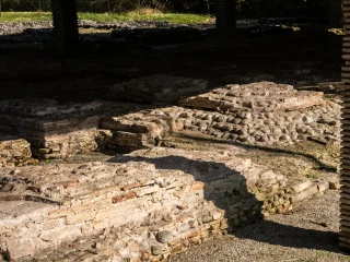 Dettaglio dell'area archeologica della villa romana di Torre con resti di strutture murarie in ciottoli e mattoni e di pavimentazioni appartenenti ad alcuni ambienti dell'edficio. In primo piano si distinguono basamenti e superfici irregolari composte da ciottoli e laterizi legati con malta. La luce solare crea zone illuminate e ombreggiate tra i resti.