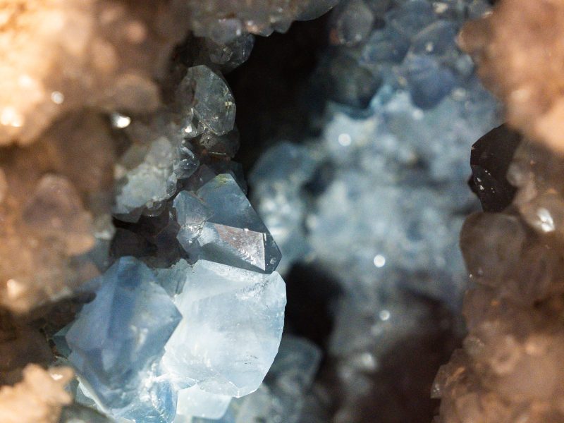 La foto in primo piano di un geode rivela cristalli sfaccettati di un azzurro brillante. I cristalli scintillanti sono circondati dalla roccia ospite ruvida e marrone chiaro. La luce illumina l'interno della cavità.