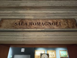 The entrance to a museum gallery featuring a stone lintel bearing the inscription “Sala Romagnoli”, above a door leading to an exhibition space.
