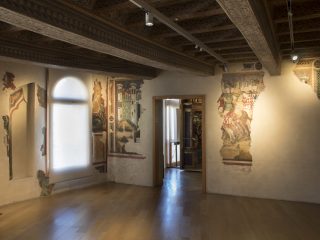 Interno di una sala museale di palazzo Ricchieri, con pavimenti in legno e soffitto a cassettoni e travature decorato. Sulle pareti chiare sono visibili frammenti di affreschi medievali che ritraggono un castello e scene di cavalieri. Una finestra ad arco sulla sinistra è coperta da una tenda bianca che diffonde la luce naturale. La scena racconta l'assedio di Pamplona.