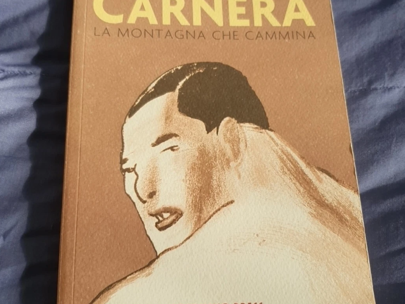 Copertina del libro "Carnera: La montagna che cammina" di Davide Toffolo, edito da Coconino Press - Fandango. L'illustrazione, realizzata con uno stile a tratti di matita o carboncino su sfondo marrone terra, ritrae il volto e le spalle massicce del pugile Primo Carnera che guarda sopra la spalla. Il titolo "CARNERA" è scritto in grandi lettere gialle nella parte superiore. Il libro è appoggiato su una superficie di tessuto blu stropicciato.