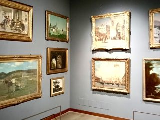 Una veduta d'angolo di una sala espositiva di una galleria d'arte o di un museo, con pareti grigio-azzurre su cui sono appesi diversi dipinti ad olio incorniciati in oro. Le opere, di diverse dimensioni, sono disposte in modo fitto e mostrano principalmente paesaggi, scene urbane e vedute classiche in stile ottocentesco o primo novecentesco. Sulla parete di sinistra risaltano un grande paesaggio rurale con un carro e una scena di mercato vivace. Sulla parete frontale, spiccano due vedute architettoniche di piazze monumentali inondate di luce solare. Le cornici sono elaborate e dorate, riflettendo la luce soffusa dei faretti della galleria. In basso, si notano i sottili cavi metallici di protezione per i visitatori e un battiscopa in legno scuro lungo il pavimento chiaro.