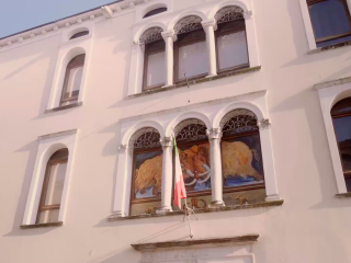 Facciata esterna di Palazzo Amalteo in stile veneziano. L'edificio, color crema, presenta eleganti trifore con archi a tutto sesto e colonnine chiare. Lo spazio della trifora centrale ospita una grande illustrazione con due mammut duellanti. Il mammut è simbolo del museo, e ricorre anche nel logo ufficiale. Una bandiera italiana sventola da un piccolo balcone in ferro battuto.