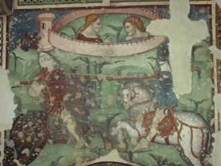 Un affresco medievale in stile gotico cortese che raffigura un duello tra due cavalieri in sella ai loro destrieri. Il cavaliere a sinistra indossa un'armatura scura e una tunica rossa, mentre quello a destra cavalca un cavallo bianco finemente bardato. Entrambi impugnano lunghe lance. Nella parte superiore, all'interno di una cinta muraria con una torre, due figure osservano la scena. Si tratta di Tristano e Isotta. L'opera presenta evidenti segni del tempo e mancanze di intonaco, con una vistosa lacuna a forma di croce sulla destra.