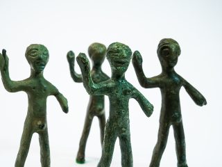 Primo piano di quattro bronzetti stilizzati con una marcata patina verde scuro. Le figure, collocate su uno sfondo bianco neutro, sono rappresentate frontalmente con il braccio destro sollevato. La texture del metallo appare rugosa e antica, e le teste presentano tratti somatici essenziali come grandi occhi incavati e bocche accennate.