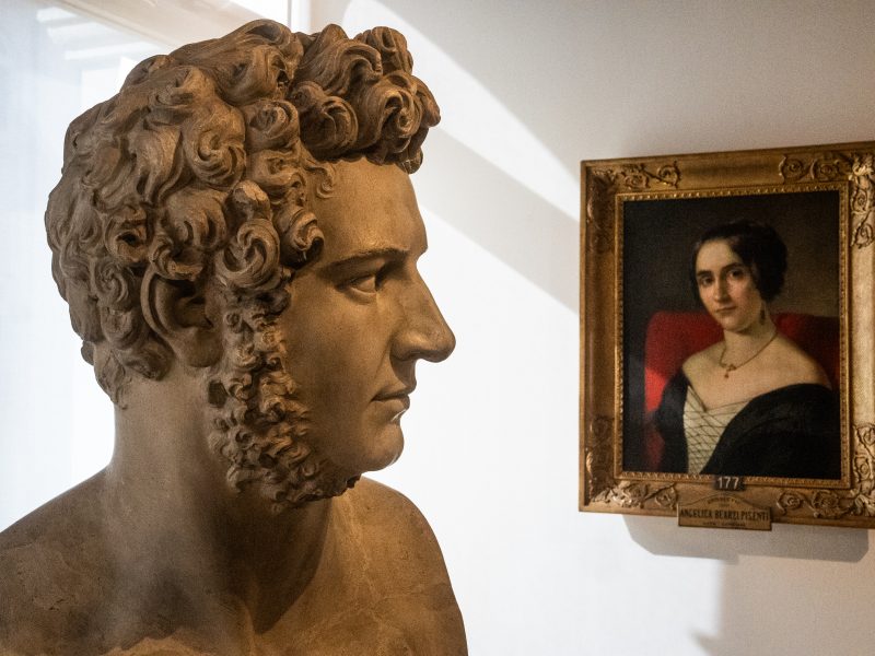 Un'inquadratura di un'esposizione museale che presenta un busto di scultura in marmo di un uomo con i capelli ricci biondo chiaro e una barba. La scultura guarda a destra. Sullo sfondo a destra c'è un dipinto a olio incorniciato in oro raffigurante una donna con i capelli scuri sollevati che indossa un abito scuro. C'è una targa di identificazione sotto il dipinto.