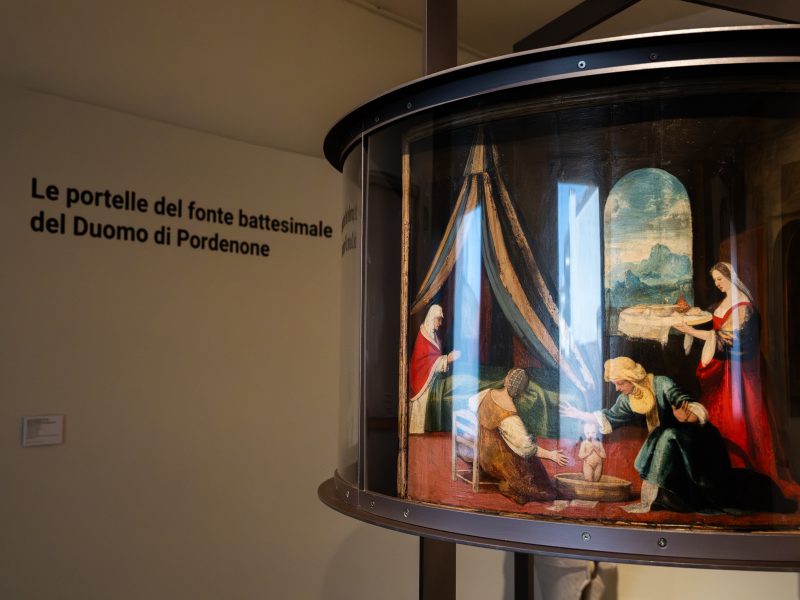 Una fotografia scattata all'interno di un museo o spazio espositivo. In primo piano, sulla destra, si trova una teca cilindrica moderna in vetro e metallo che protegge un antico dipinto su tavola. L'opera raffigura una scena d'interno con diverse figure femminili in abiti rinascimentali; una donna è a letto sotto un baldacchino, mentre altre figure si prendono cura di un neonato in primo piano. Sullo sfondo della teca, a sinistra, una parete bianca ospita una scritta nera di grandi dimensioni: "Le portelle del fonte battesimale del Duomo di Pordenone". L'illuminazione è soffusa, tipica di un ambiente museale, e mette in risalto il contrasto tra il reperto storico e l'allestimento contemporaneo.