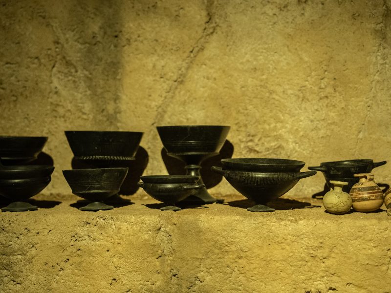 Dettaglio di alcuni vasi della collezione Micheluzzi all'interno dell'ambientazione che riproduce una tomba etrusca. A sinistra e al centro predominano coppe e calici neri in bucchero, caratterizzati da forme eleganti con steli e anse. Sulla destra si trovano tre piccoli vasi globulari in argilla chiara, decorati con motivi bruni.