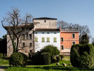 Primo piano del Castello di Torre. A sinistra si nota la parte più antica del castello, dietro a un grande albero spoglio. Al centro, una parte del complesso intonacata di bianco e dotata di balconi verdi è sovrastata dalla torre centrale; a destra una sezione rosata con balconi verdi e un terrazzo con balaustra. In primo piano, un prato verde con siepi decorative di varie forme, tra cui una grande siepe sulla destra.