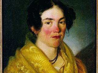 Un dipinto a olio che ritrae a mezzo busto una giovane donna rivolta leggermente verso lo spettatore. Il volto è caratterizzato da guance e naso arrossati, occhi scuri e un'espressione serena. I capelli, neri e mossi, sono raccolti lasciando scoperte le orecchie, dove si nota un piccolo orecchino a cerchio dorato. La donna indossa uno scialle giallo ocra luminoso, riccamente lavorato con motivi che ricordano piume o ricami floreali, avvolto sopra un abito scuro di cui si intravede la scollatura bianca. Al collo porta una collana a doppio giro di piccole perle scure. Lo sfondo è scuro e uniforme, quasi nero, il che fa risaltare la luminosità dell'incarnato e il giallo acceso del vestiario. L'opera è racchiusa in una sottile cornice di legno scuro e, nel bordo inferiore, si scorge parzialmente un numero di catalogazione. Lo stile pittorico è realistico, tipico della ritrattistica del XIX secolo.
