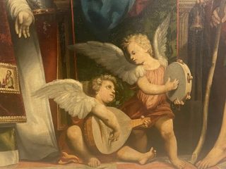 Dettaglio di un dipinto rinascimentale raffigurante due angeli alati che suonano strumenti musicali ai piedi del trono della Madonna. Si tratta della pala d’altare di Giovanni Antonio de’ Sacchis detto il Pordenone proveniente dalla Chiesa parrocchiale dei Santi Ilario e Taziano, nella frazione Torre. L'angelo a sinistra è seduto a terra e suona un liuto; indossa una tunica arancione. L'angelo a destra è in piedi e suona un tamburello; indossa una tunica rosa-rossa. Entrambi hanno grandi ali bianche sfumate. Sullo sfondo si intravedono le ricche vesti rosse e dorate e il manto blu scuro delle figure centrali di santi e della Madonna.