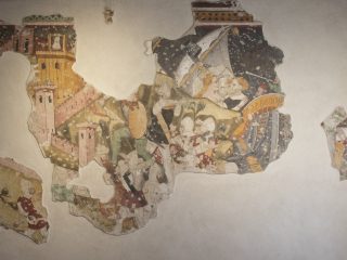 Frammenti di affresco medievale raffiguranti scene della leggenda di Tristano e Isotta, con personaggi, architetture e decorazioni policrome su fondo chiaro.