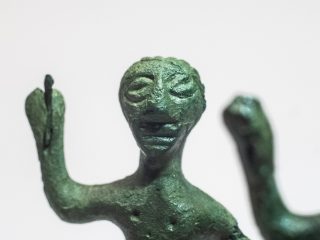 Macrofotografia del volto di un bronzetto con patina verde scuro. I tratti somatici sono resi in modo essenziale: grandi occhi a mandorla incavati, naso pronunciato e una fessura per la bocca. Sopra il labbro sono rappresentati forse baffi stilizzati. La testa è incorniciata da una capigliatura portata all'indietro. Il metallo presenta una texture granulosa e irregolare. Sullo sfondo sfocato si intravede la mano destra sollevata che impugna un frammento dell'originale lancia.