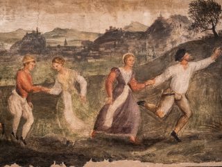 Un dettaglio di un affresco rinascimentale che ritrae due coppie impegnate in una danza o in una corsa festosa all'aperto. Le figure sono disposte in modo dinamico: A destra: Un uomo con una camicia bianca e un berretto nero corre con slancio in avanti, indicando qualcosa con il braccio teso, mentre tiene per mano una donna in un abito viola e arancione che lo segue sorridendo. A sinistra: Una seconda coppia si tiene per mano con un gesto più pacato; l'uomo indossa una giubba rossa e pantaloni chiari a sbuffo, mentre la donna indossa un abito lungo di colore verde pallido. Sullo sfondo si estende un ampio paesaggio collinare con castelli e borghi fortificati arroccati sulle cime, resi con tonalità terrose e soffuse. La superficie del dipinto mostra i segni del tempo, con piccole crepe e una texture murale che conferisce all'opera un aspetto storico e materico. I colori dominanti sono l'ocra, il terra di Siena e il verde oliva.