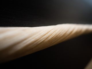 Un'inquadratura macro e ravvicinata mostra un dettaglio della superficie del dente di narvalo, mettendo in risalto la sua particolare conformazione naturale. La sezione del dente attraversa l'immagine in diagonale. La messa a fuoco è selettiva e concentrata sulla parte centrale, rivelando una fitta trama di scanalature e rilievi a spirale che corrono lungo l'avorio. Il materiale di cui è formato il dente è di color crema e contrasta nettamente con lo sfondo sfocato, molto scuro, facendo quasi "emergere" la zanna dal buio. La profondità di campo ridotta enfatizza la tridimensionalità della spirale, trasformando un reperto biologico in un dettaglio quasi astratto e scultoreo.