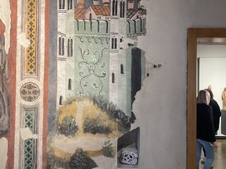 Frammento di affresco medievale su una parete museale che raffigura un castello stilizzato con alte torri bianche, merlature e tetti rossi. La facciata del castello è decorata con motivi a viticci bianchi su fondo verde. Sulla sinistra è presente una cornice decorativa verticale con motivi geometrici e floreali. A destra, una porta aperta lascia intravedere dei visitatori in una sala adiacente. La scena racconta l'assedio di Pamplona: in particolare si vede la città con la cinta murata.