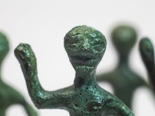 Primo piano ravvicinato del volto di una delle statuetta in bronzo con patina verde da Praturlone. I lineamenti sono stilizzati, con ampie orbite oculari e pupille rappresentate da linee orizzontali. La bocca è segnata da un'incisione orizzontale, mentre una serie di liee verticali indicano la presenza di una barba. Sullo sfondo, fuori fuoco, si intravedono il braccio sollevato della figura e le sagome di altre statuette simili.