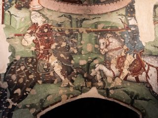 Dettaglio di affresco medievale della leggenda di Tristano e Isotta raffigurante uno scontro cavalleresco con figure armate e cavalli su fondo verde decorato.