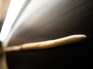 Un'inquadratura ravvicinata a fuoco selettivo della punta affusolata del dente di narvalo (la zanna) visto nell'immagine precedente, posizionato orizzontalmente ed emergente verso destra. I dettagli principali includono: Soggetto: La punta è la parte più nitida e ha una colorazione crema-avorio liscia e calda. Procedendo verso sinistra, la zanna si allontana dal piano focale, diventando progressivamente sfocata, rivelando una texture spiraleggiante e più ruvida con sfumature dorate e macchie naturali. Composizione: La zanna attraversa diagonalmente l'immagine dal basso a sinistra verso l'alto a destra, creando una linea d'azione dinamica. La profondità di campo molto ridotta focalizza l'attenzione sulla solidità della punta. Sfondo: Lo sfondo è diviso in due zone chiaramente contrastanti. La parte superiore e inferiore sono occupate dal supporto espositivo ovale nero, che appare scuro e completamente sfocato. A sinistra, una fonte di luce bianca intensa e brillante crea un bagliore diffuso, quasi un'aurora, che sfoca i contorni della zanna. L'illuminazione mette in risalto la forma appuntita e la solidità della zanna, catturando un dettaglio tipico della tassidermia museale di alta qualità.